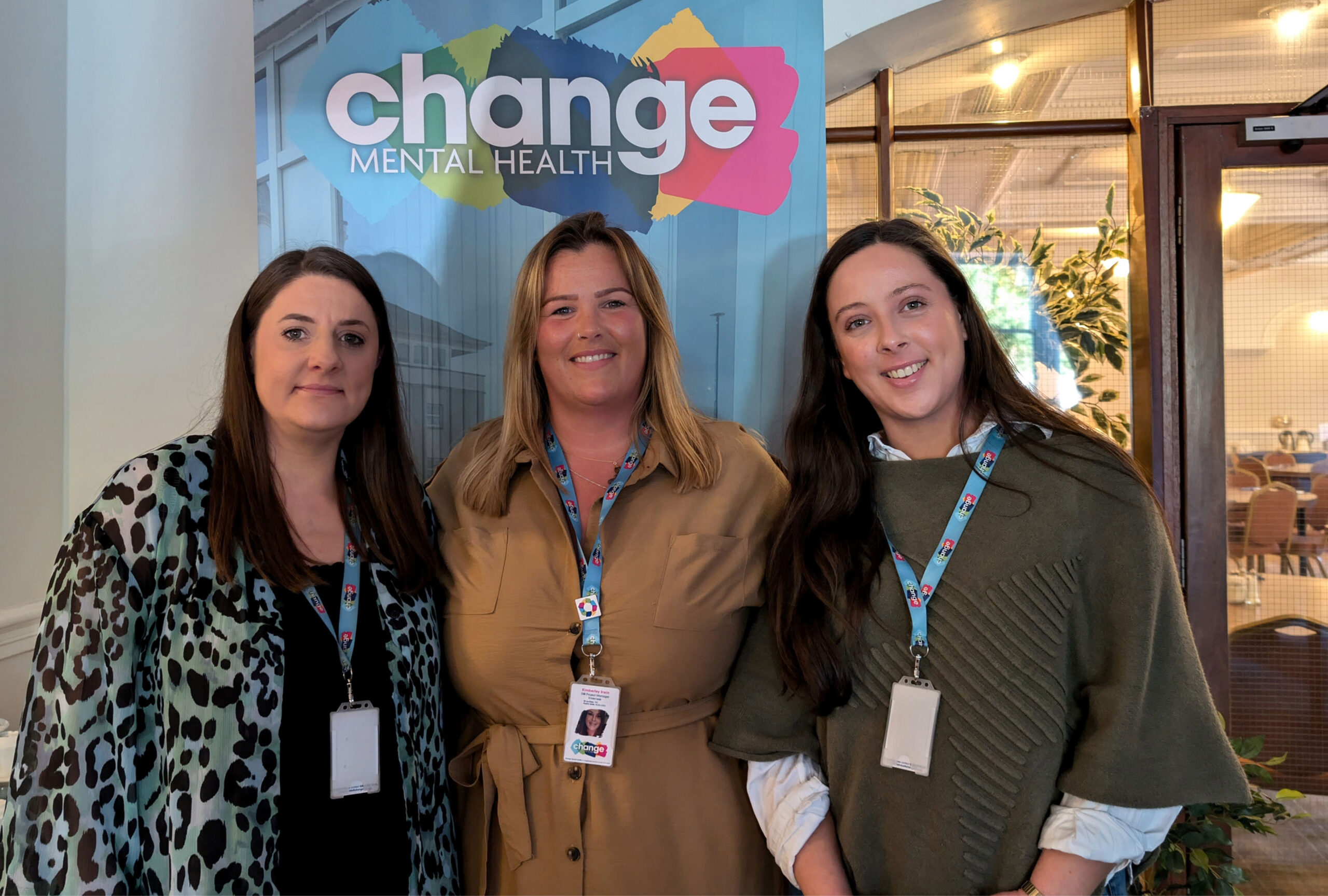 Three women working for DBI stand together indoors, smiling, wearing lanyards in front of a colorful 'Change Mental Health' sign.