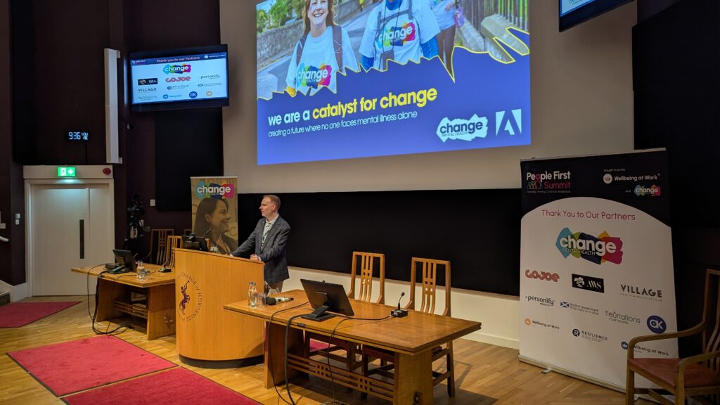 Speaker at a wooden podium in a conference room, large slide behind him reads 'we are a catalyst for change' with Change Mental Health branding.