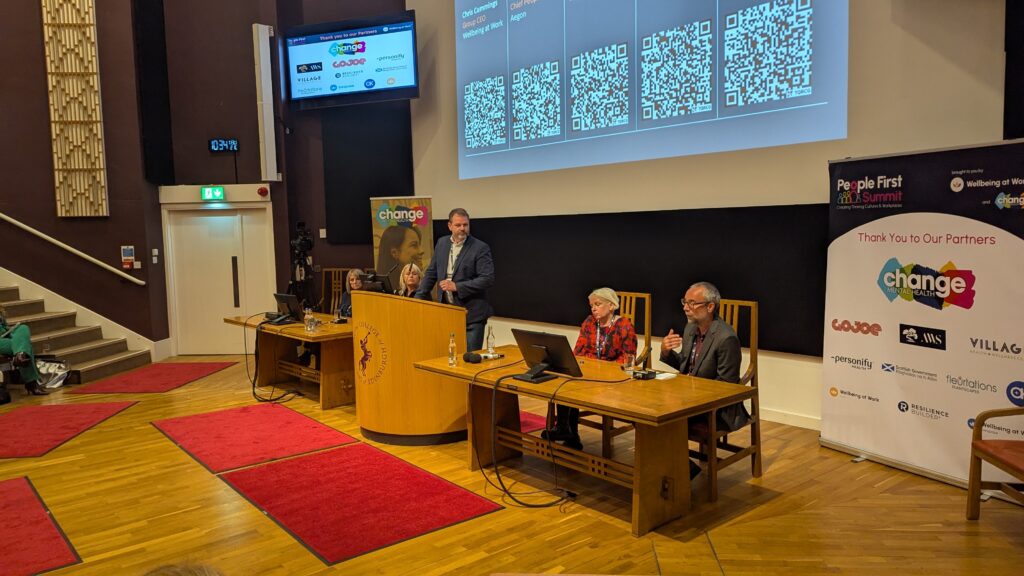 Speaker at podium addressing a panel of four people at a conference table in a lecture hall, with a large screen displaying QR codes behind them.