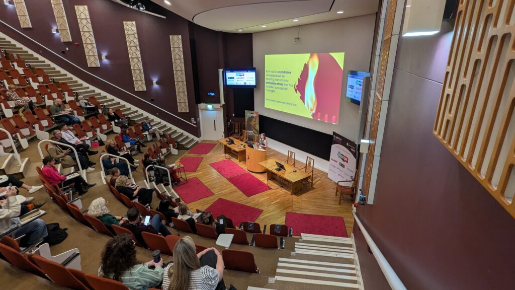 Auditorium with curved red seats and attendees watching a presentation on stage with a large yellow slide projected on screen.