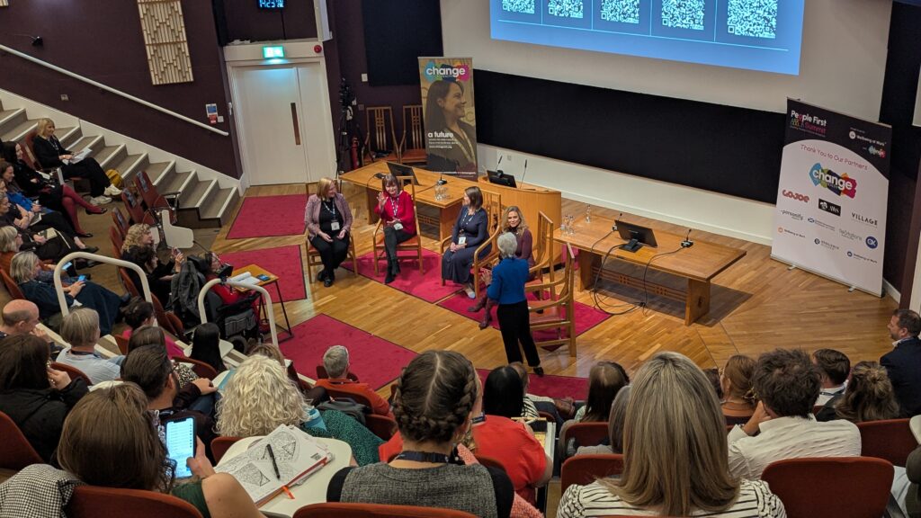 Panel discussion in a university-style auditorium with four women seated on stage and a banner nearby.