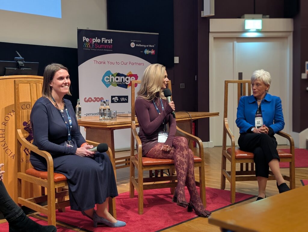 Panel discussion with three women seated on wooden chairs in a conference room, banner reading 'Change Mental Health' behind them, microphones and water bottles on the table nearby.