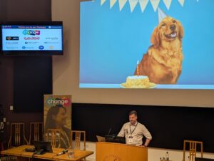 Photo from 2025' People First Summit featuring Jonnny Cobbold talking about Happiness at Work. Presenter at a podium in a conference hall; a large blue screen shows a golden retriever with a birthday cake, while a smaller screen to the left displays partner logos and 'Thank you to our Partners' banner.