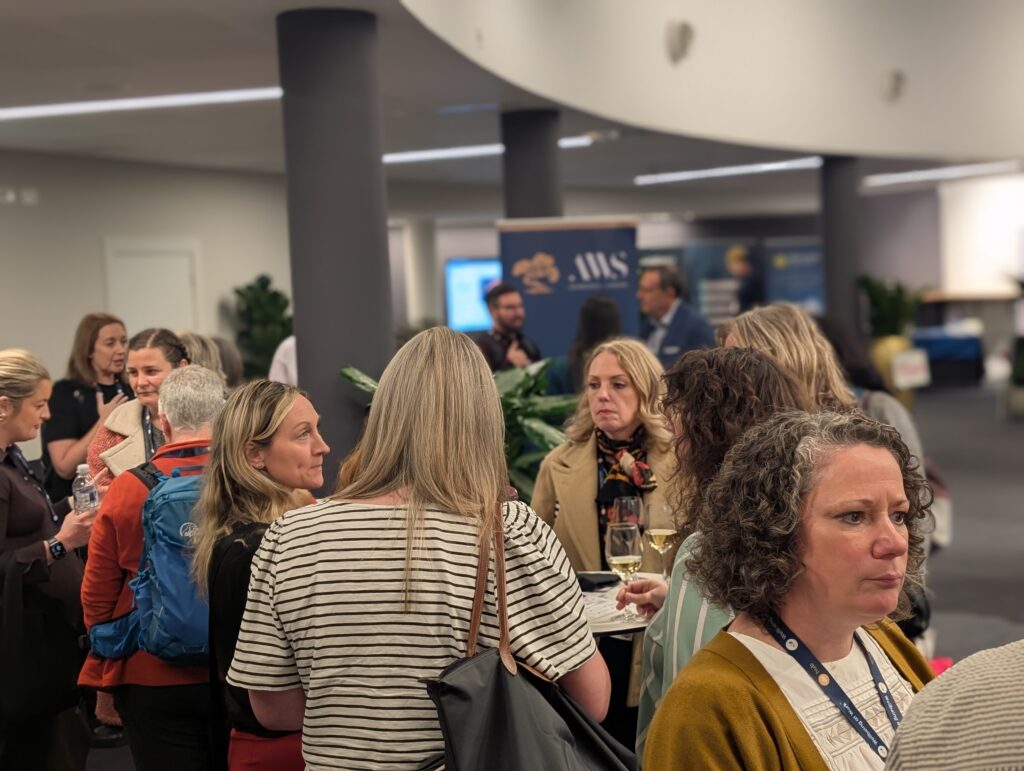 Crowd of professionals mingling at an indoor conference, some holding wine glasses, with banners in the background and lanyards around their necks.