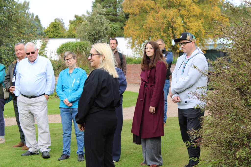 A group of people standing outdoors in a garden, wearing jackets and casual clothing, with trees showing autumn colors in the background.