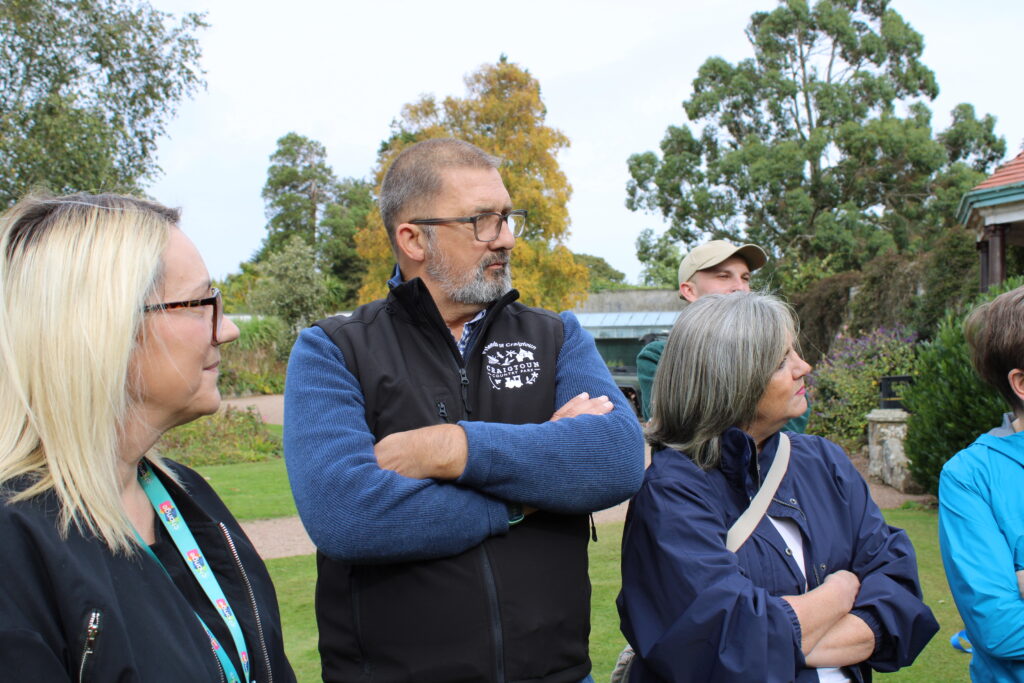 Group of adults outdoors in a park, man with glasses and beard at center standing with arms crossed as others look on.
