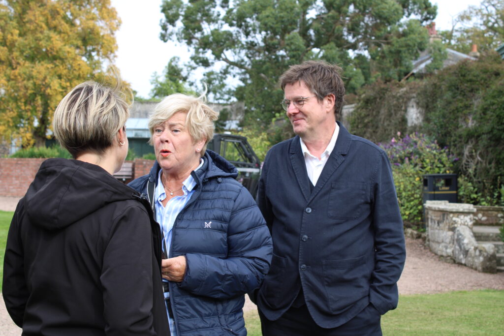 Three adults chatting outdoors in a garden: a blonde woman in a blue quilted jacket at center, a man in a dark suit to her right, and another person with a black jacket on the left (rear view).