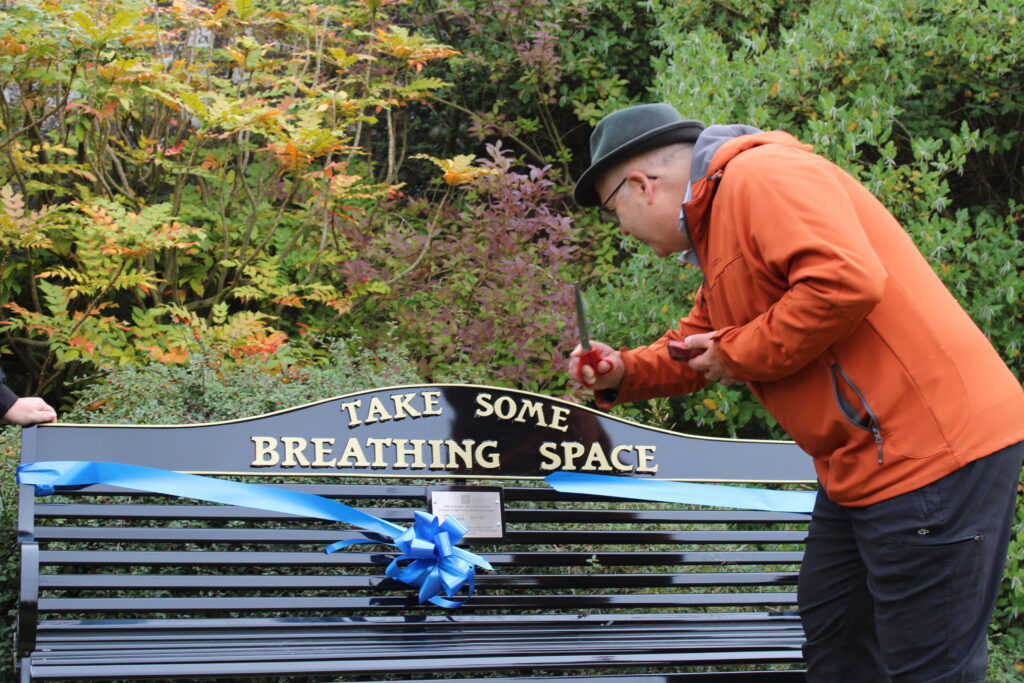 Man in an orange jacket and black hat cuts a blue ribbon in front of a bench with a sign reading ‘Take Some Breathing Space.’