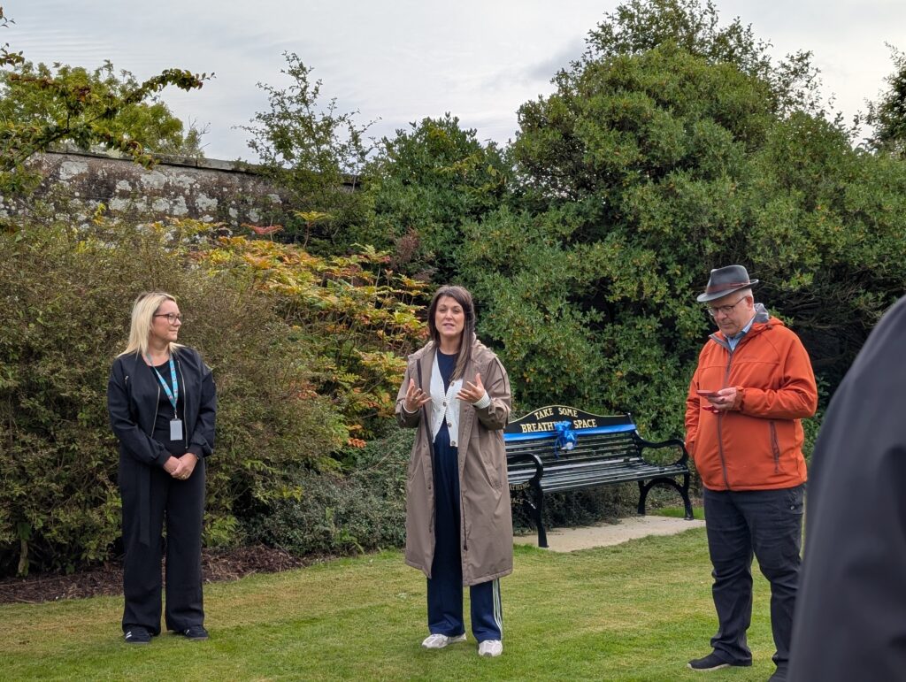 Three people stand outdoors in a garden, with one woman speaking in a beige coat while others listen. A bench and greenery are behind them.