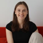 Ffion Hargood smiling at the camera, sitting on a red couch in a black top and beige pants.