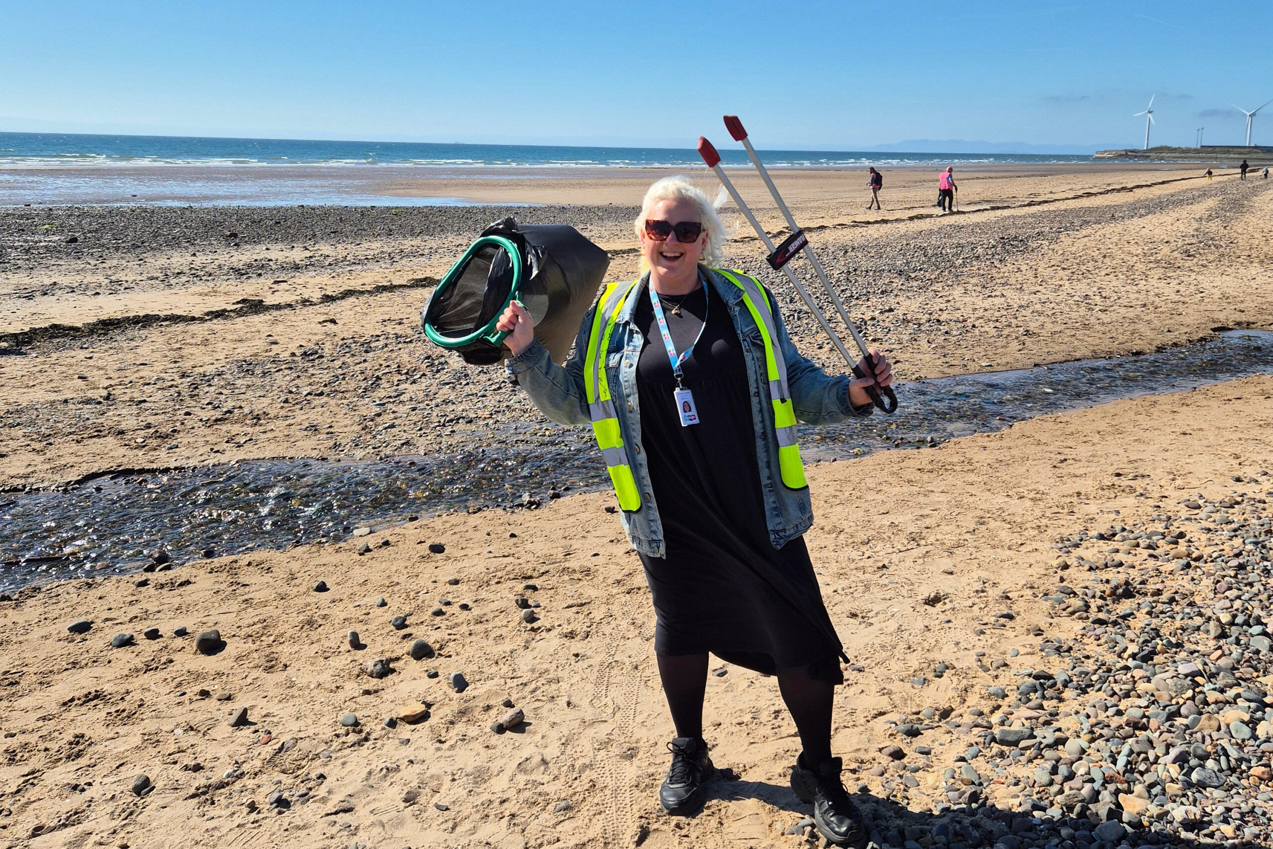 Smiling woman - Jaz Lacey-Campbell, the co-author of the Climate strategy - in a high-visibility vest and denim jacket on a pebbly beach during the litter picking