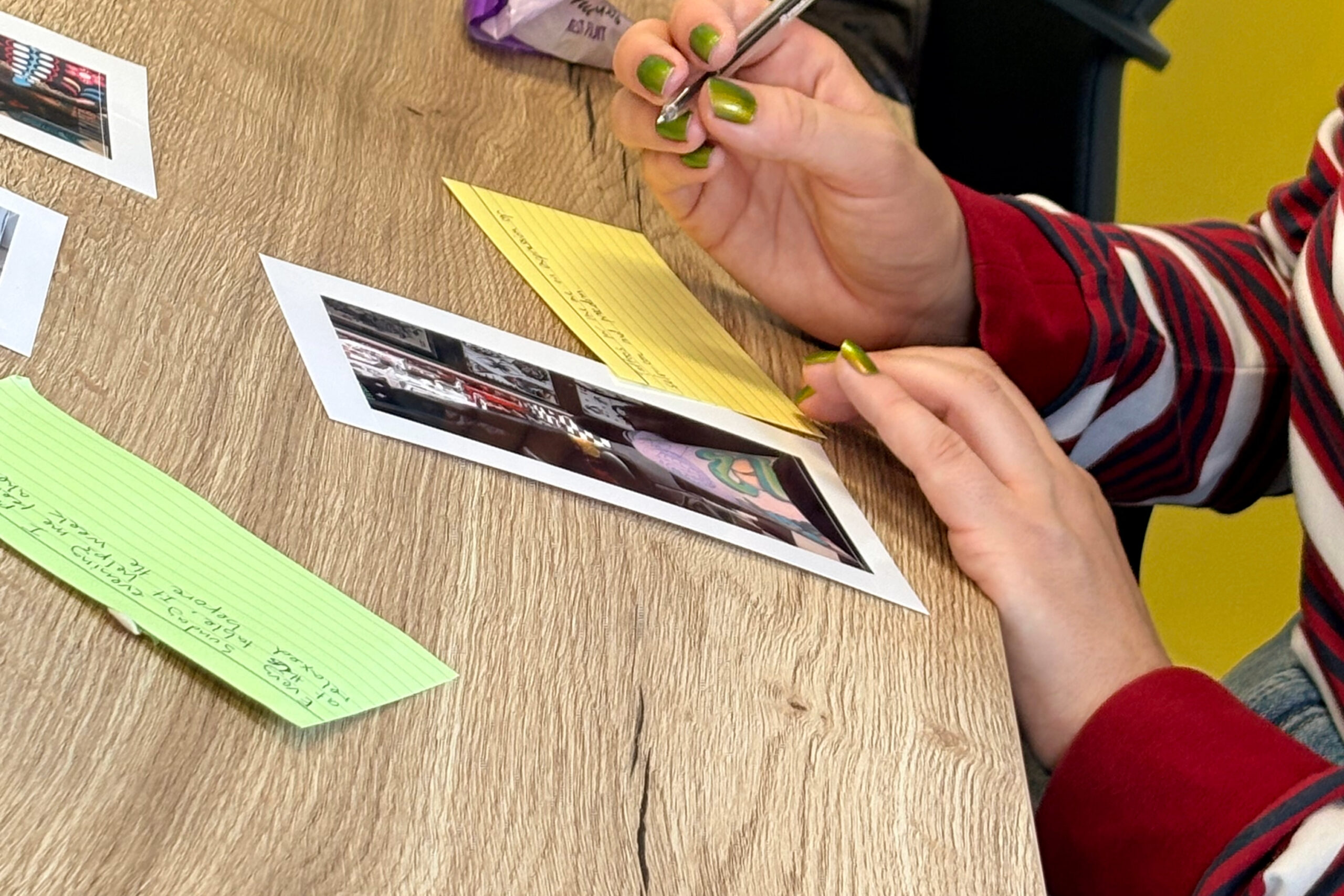 Person with green nails writes on a yellow lined notebook at a wooden table, with photo strips nearby.