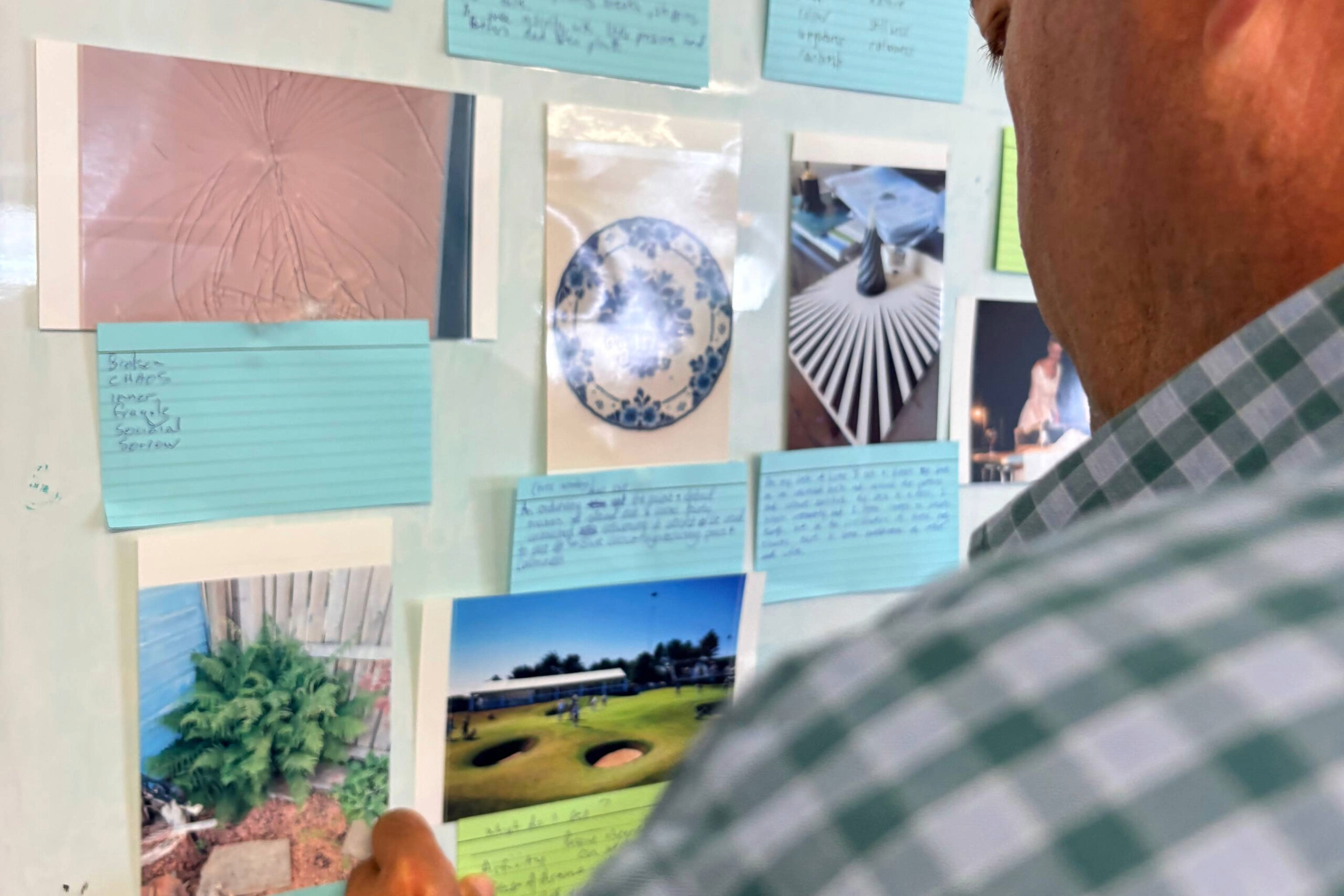 Person in a green-and-white checkered shirt looking at a wall covered with blue sticky notes and photos.