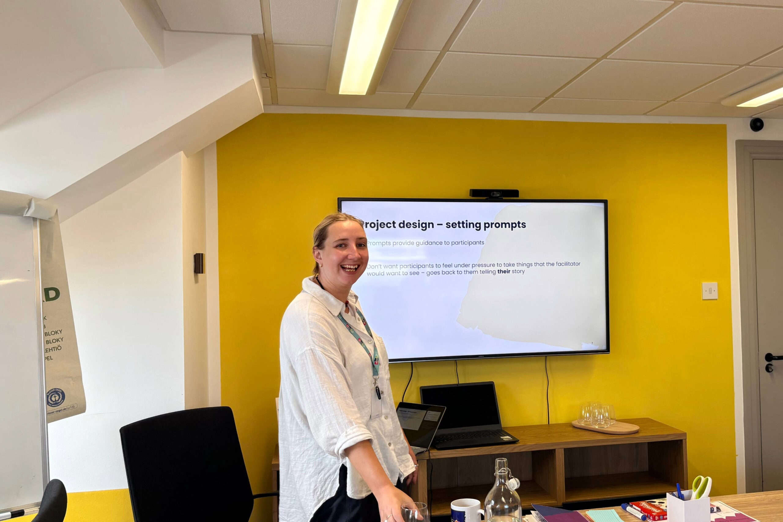 Smiling woman presenting in a meeting room with a bright yellow wall and a large screen showing a slide about project design – setting prompts.