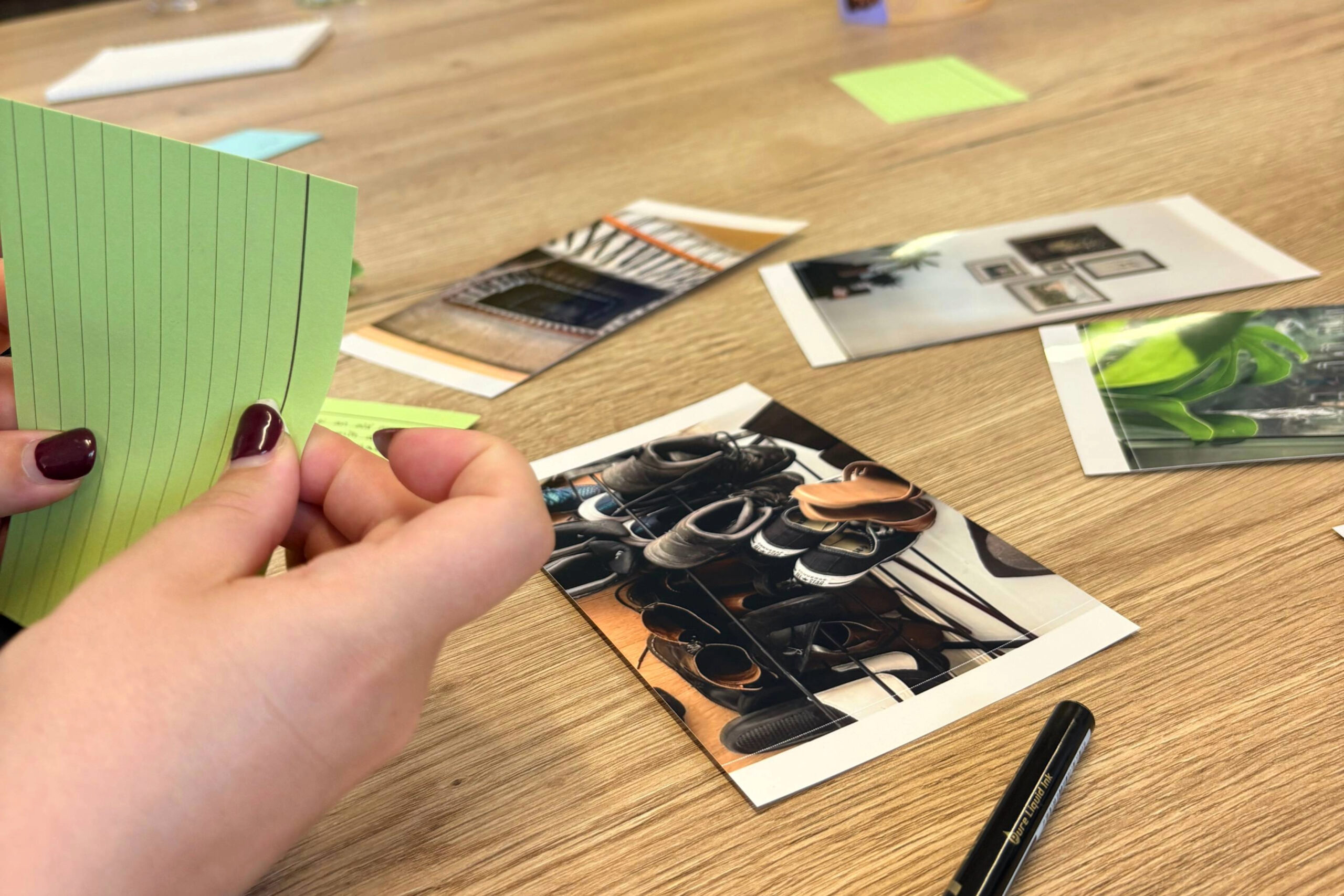 Close-up of hands holding a green lined notecard on a wooden table with scattered photos and magazines nearby.